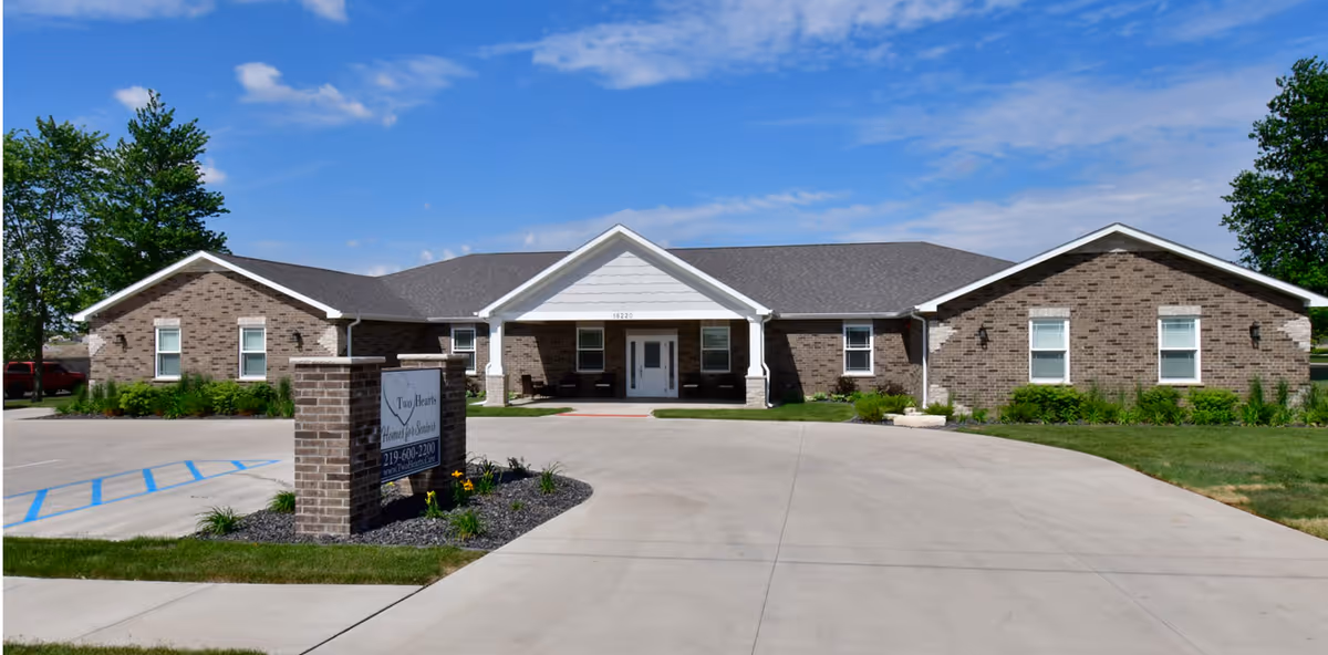 Front view of a one-story brick senior living building with a covered entrance, driveway, landscaping, and a sign reading 'Two Hearts Homes For Seniors'.