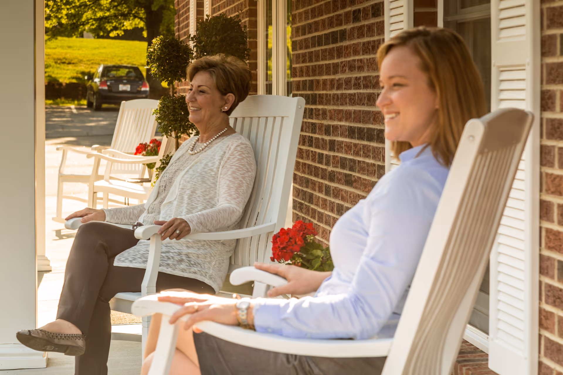 Two women sitting and relaxing on white rocking chairs on a porch outside a brick building, with red flowers and greenery nearby.