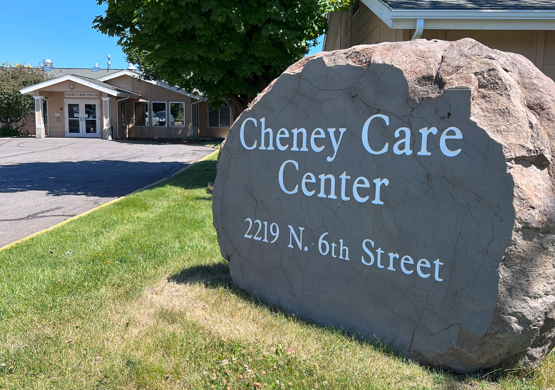 Large rock with the words 'Cheney Care Center 2219 N. 6th Street' engraved on it, situated on a grassy area with a tree behind it. In the background, there is a single-story building with a sign above the entrance that reads 'Cheney Care Center'.
