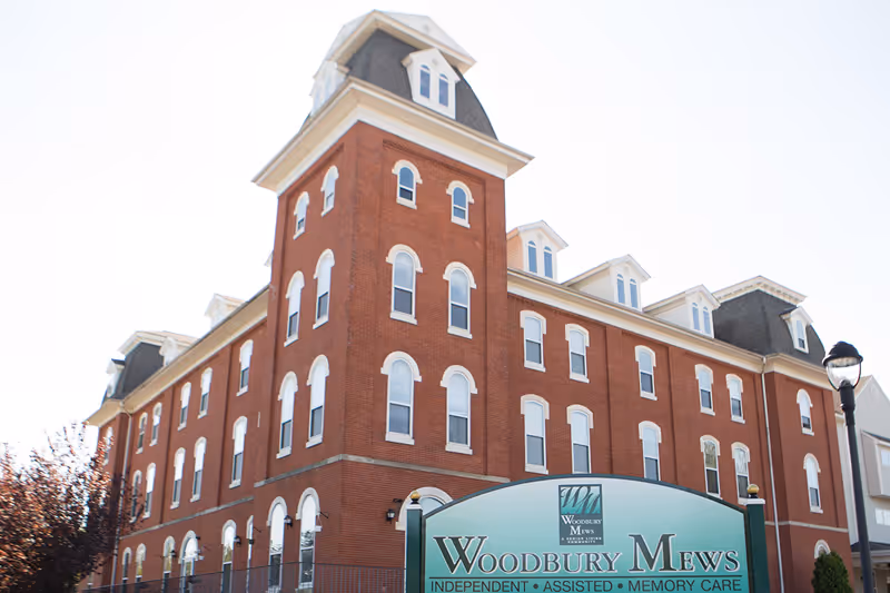 Exterior view of a large, multi-story red brick building with white-trimmed windows and a mansard roof, identified as Woodbury Mews senior living facility by a green sign in front indicating independent, assisted, and memory care services.
