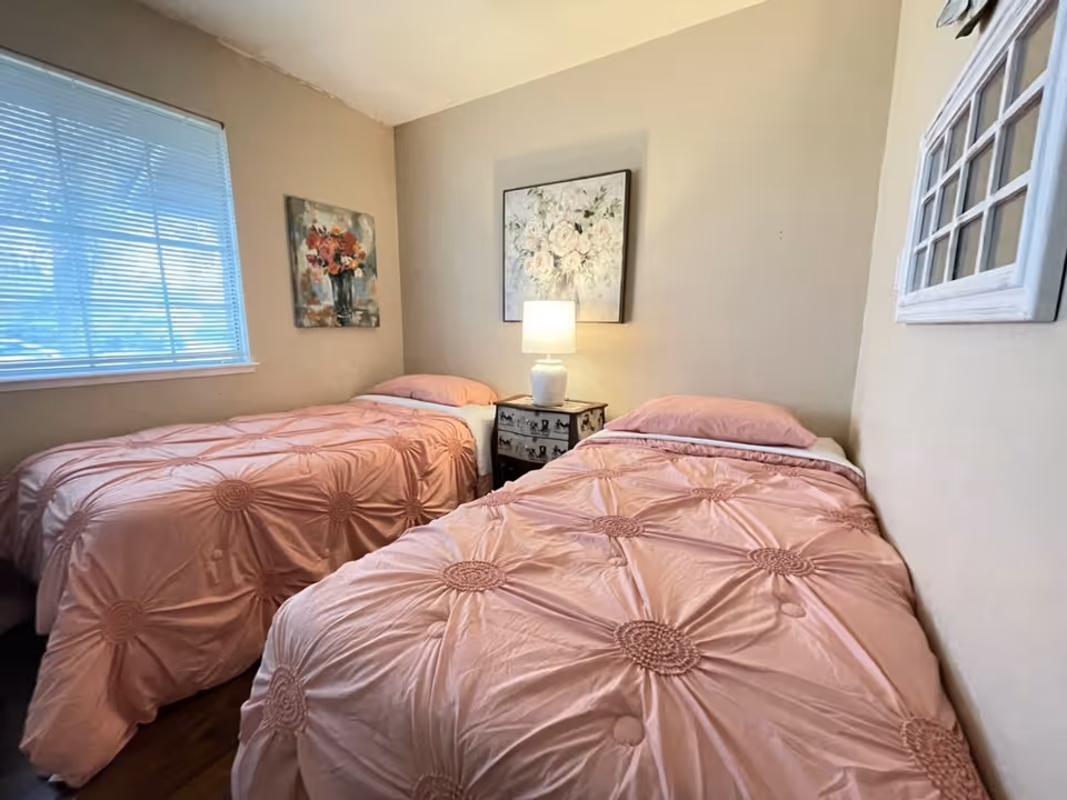 Small bedroom with two twin beds in pink comforters, a nightstand with a lamp between them and floral wall art.