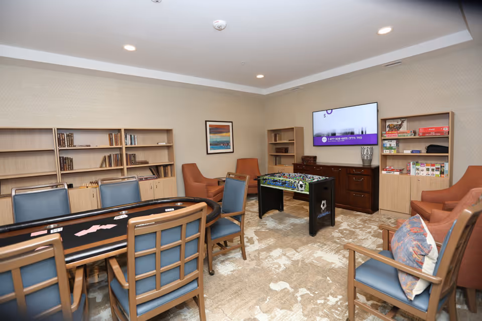 A senior living common room with a poker table, foosball table, TV, bookshelves, and multiple chairs.