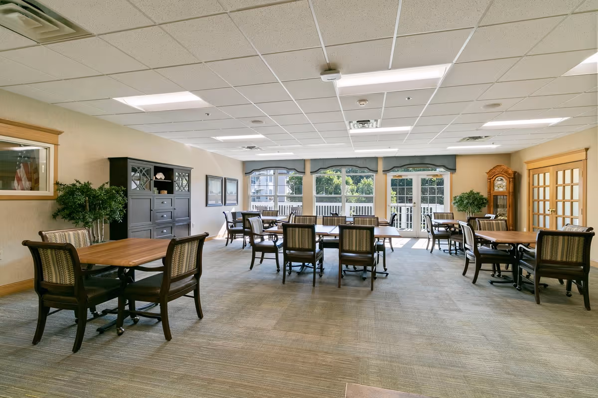 A spacious, well-lit dining room with multiple wooden tables and cushioned chairs arranged neatly. Large windows and glass doors at the far end allow natural light to fill the room. The room features a grandfather clock, a dark wooden cabinet, framed pictures on the wall, and potted plants for decoration.