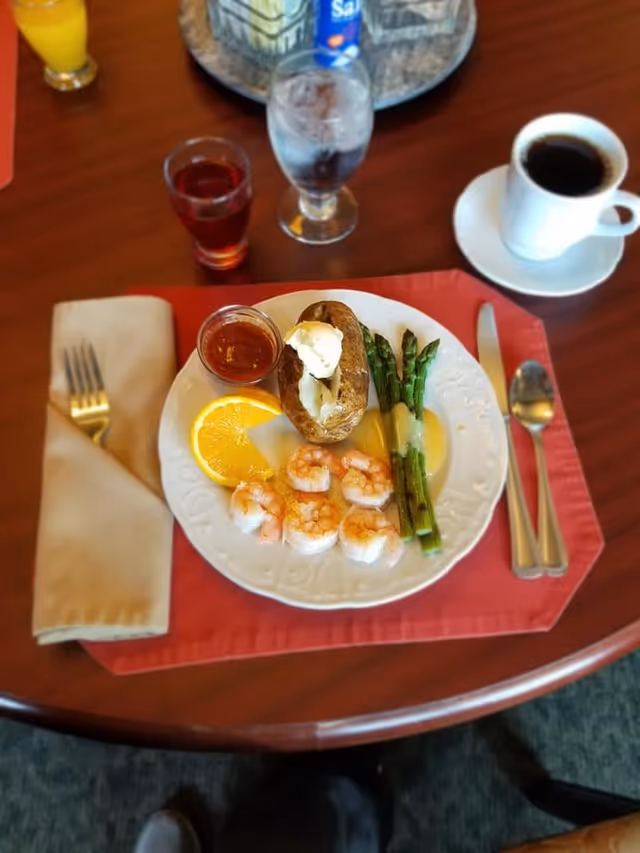 A meal served on a white plate with shrimp, grilled asparagus, a baked potato topped with butter, a slice of orange, and a small container of sauce. The plate is on a red placemat on a wooden table, with a fork and napkin on the left and a knife and spoon on the right. There is a glass of water, a cup of black coffee on a saucer, and a glass of red beverage also on the table.