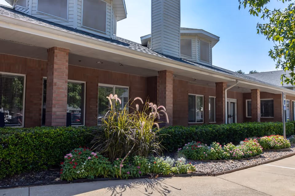 Exterior view of a brick building with multiple windows and a covered walkway supported by brick columns. There are well-maintained bushes and flowering plants along the front of the building, and a clear blue sky is visible above.