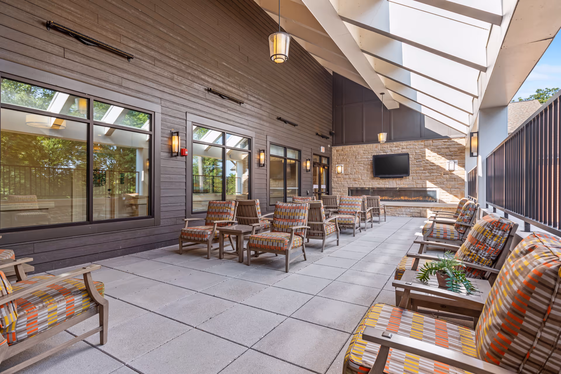 Covered outdoor patio area with multiple cushioned wooden chairs arranged in rows facing a stone wall with a mounted flat-screen TV and a long fireplace beneath it. The space has large windows on one side and a slanted roof with beams allowing natural light to enter.