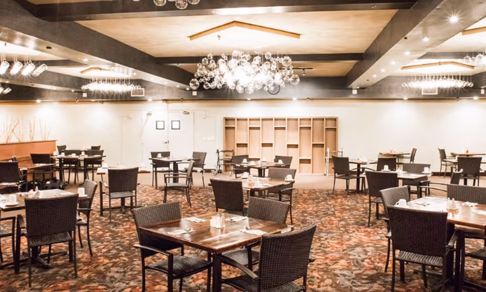 Spacious empty dining room with multiple square wooden tables and woven chairs under decorative chandeliers.