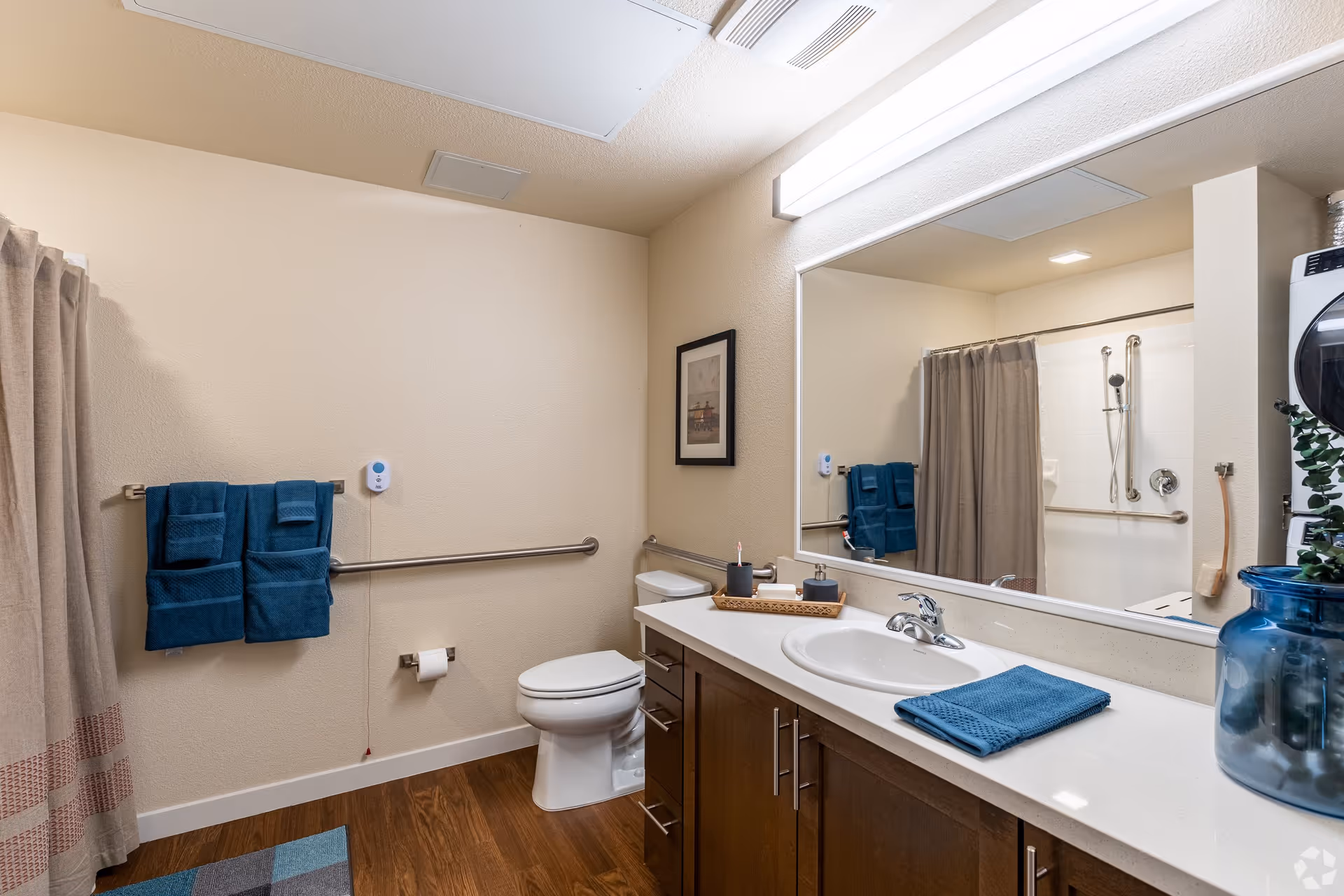 A clean and modern bathroom with beige walls and wood flooring. The bathroom features a white toilet, a large mirror above a white countertop with a sink, and dark wood cabinets. Blue towels hang on a metal towel rack next to the toilet, and a blue towel is folded on the countertop. A shower with a beige curtain and grab bars is visible in the reflection of the mirror. There is a framed picture on the wall and a blue glass vase with greenery on the counter.