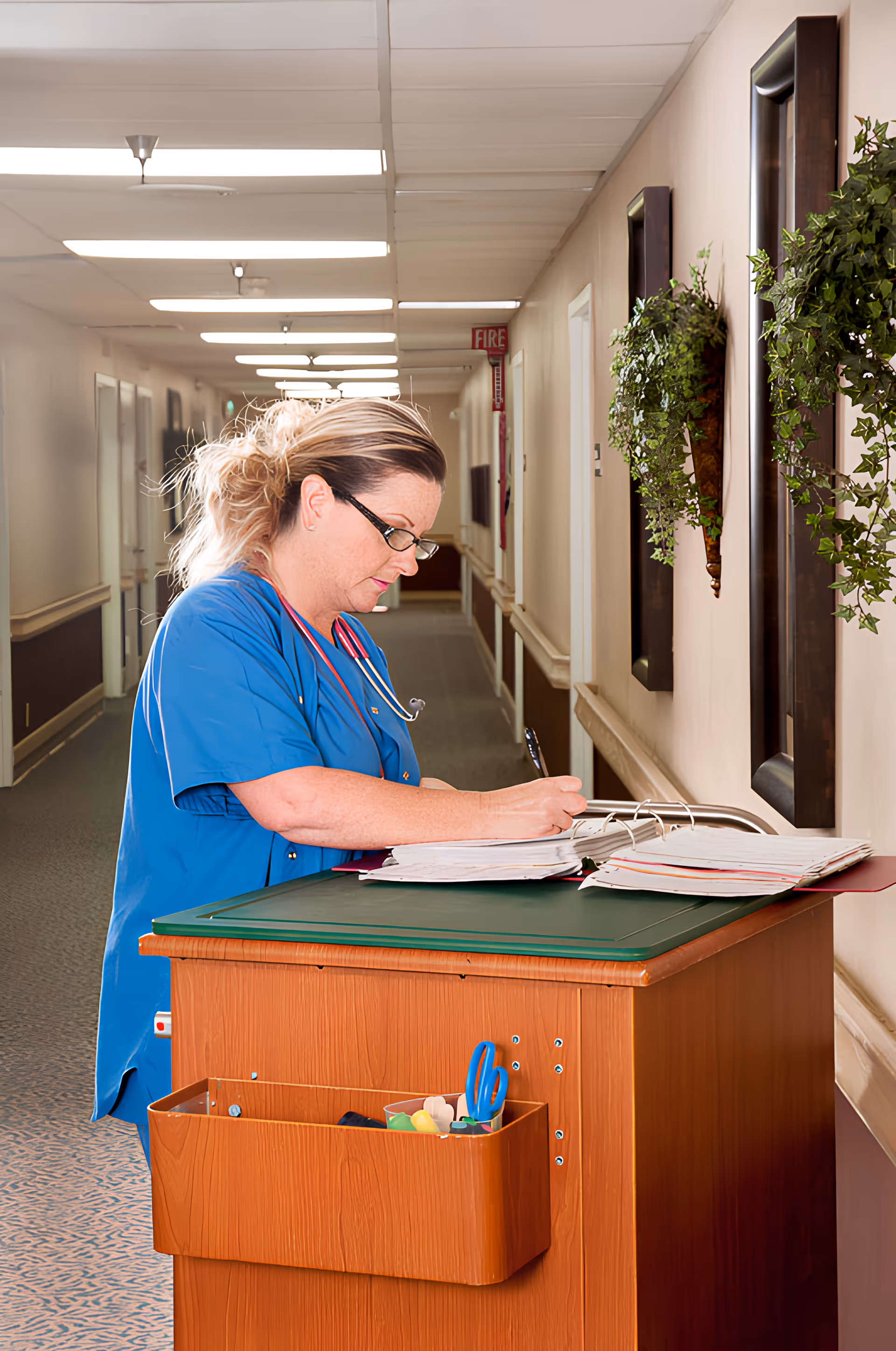 A healthcare worker in blue scrubs with a stethoscope around her neck is writing in a large binder on a wooden cart in a well-lit hallway decorated with green plants on the walls.