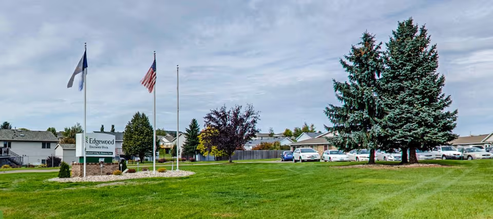 A wide grassy lawn with three flagpoles, two of which have flags raised. A sign reads 'Edgewood Bismarck on Dominion' with additional text about assisted living and memory care. Several houses and parked cars are visible in the background under a cloudy sky.