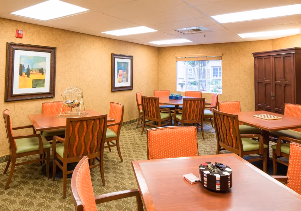 A well-lit room with multiple wooden tables and chairs with orange and green cushions. The room has beige walls with two framed paintings, a window showing an outdoor view, and a large wooden cabinet. Various game items like poker chips, playing cards, and a bingo cage are placed on the tables.