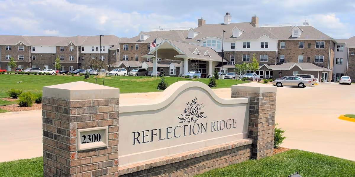 Exterior view of Reflection Ridge, a large multi-story senior living facility with a brick and beige facade. In the foreground is a decorative stone sign with the facility's name and address number 2300. Several cars are parked in front of the building, and there is a well-maintained lawn and landscaping around the entrance.
