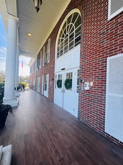 Wide exterior view of a brick building with white double doors decorated with green wreaths, large white columns supporting a covered porch, and an American flag visible in the background.