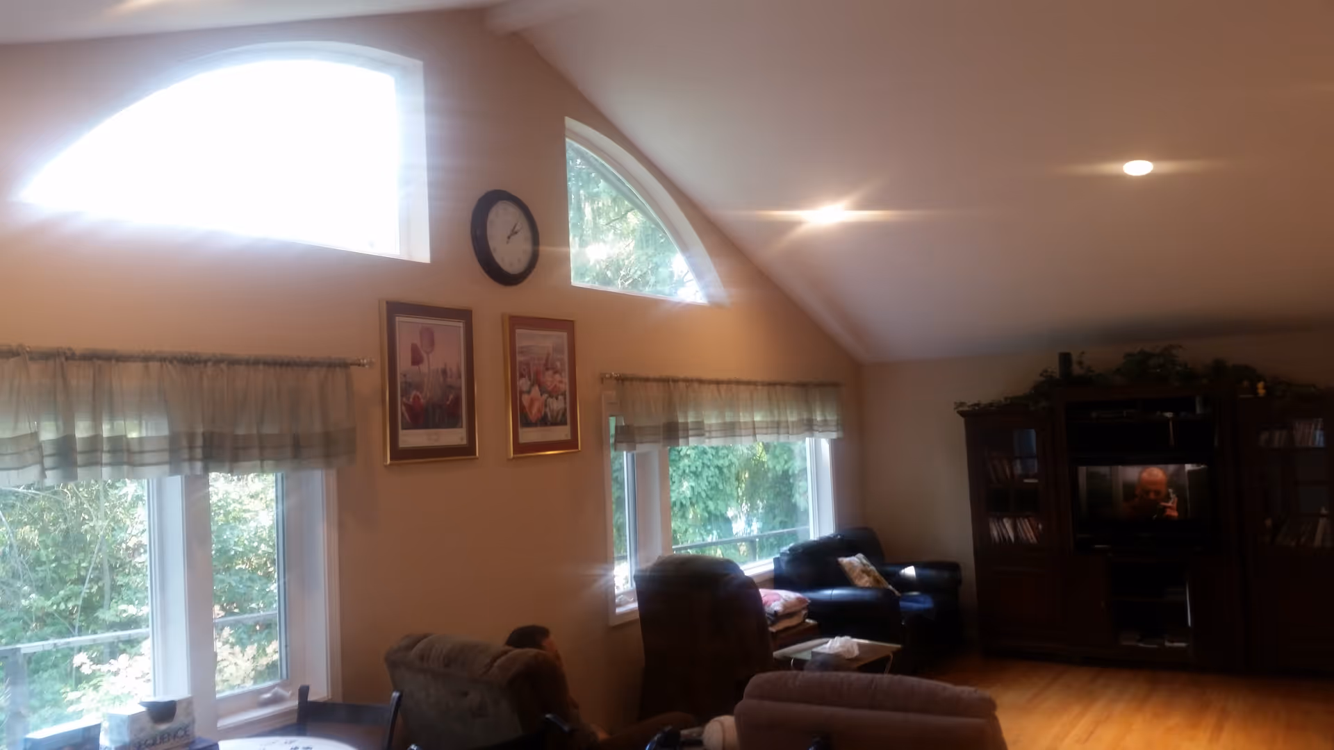 A cozy living room with large windows covered by sheer curtains, allowing natural light to brighten the space. The room features several comfortable chairs and a dark leather sofa arranged around a wooden coffee table. On the far wall, there is a large wooden entertainment center with a TV and shelves filled with books and decorative items. Above the windows, there are two framed pictures and a round wall clock. The room has a sloped ceiling with recessed lighting and wooden flooring.