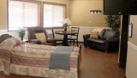 A cozy senior living room with a bed, sofa, round table, chairs, and large windows.