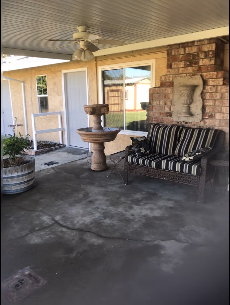 Covered outdoor patio area with a ceiling fan, a striped cushioned wicker loveseat, a decorative stone fountain, a potted plant, and a brick wall with a mounted stone urn decoration. There is a sliding glass door and a white door on the beige exterior wall of the building.