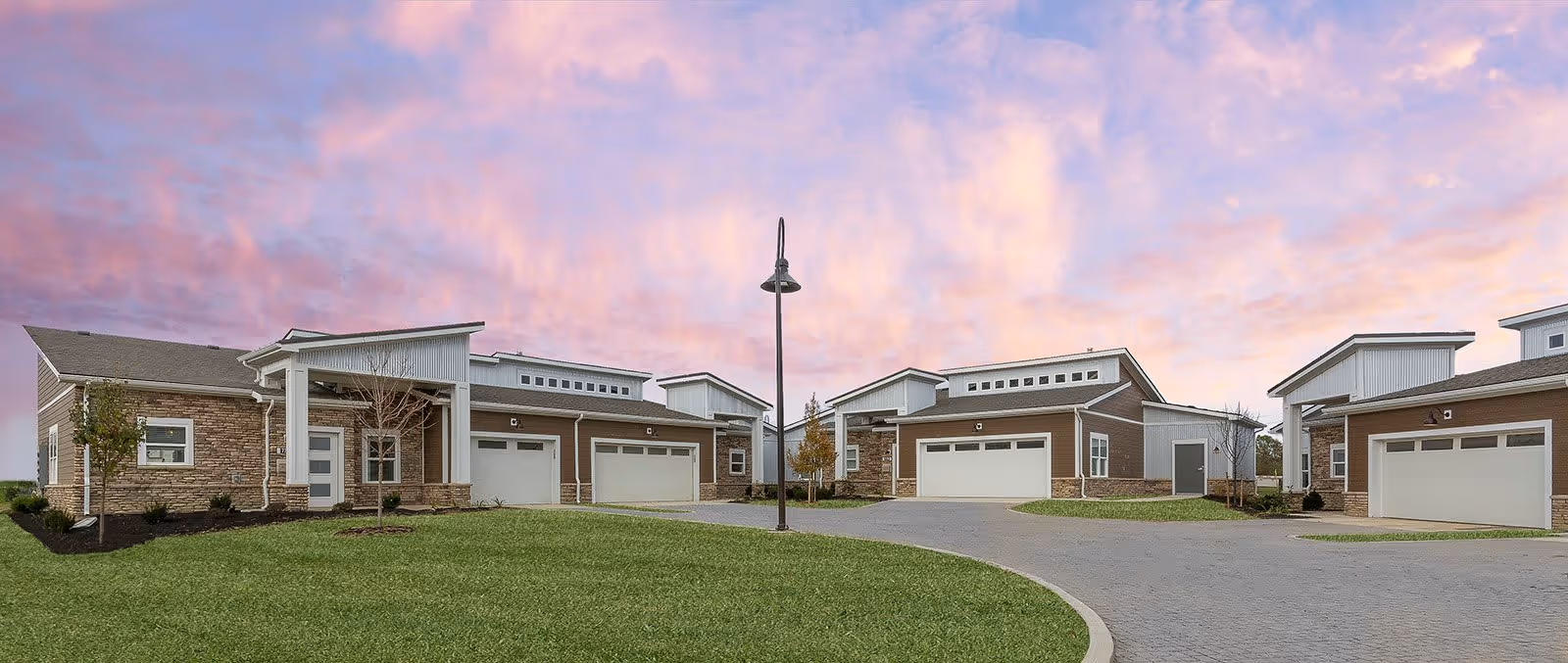 Exterior view of multiple single-story residential buildings with garages, set against a colorful sunset sky with pink and purple clouds. The buildings have a modern design with stone and wood siding, and there is a paved driveway and green lawn in the foreground.