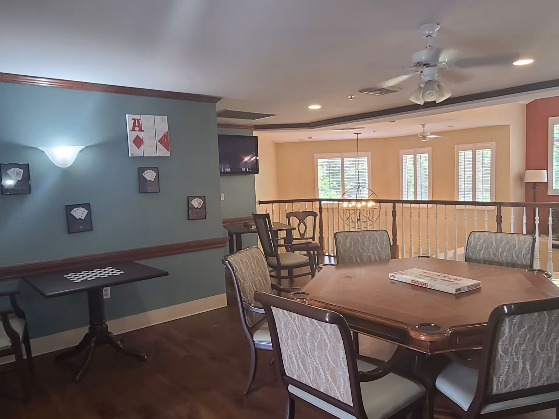 A cozy common area with a wooden card table surrounded by six upholstered chairs. A Monopoly board game is placed on the table. The room has wooden flooring and a teal accent wall decorated with framed playing cards. There is a small table with a checkerboard pattern and chairs against the wall. The area is well-lit with ceiling lights and a ceiling fan, and windows with white blinds allow natural light to enter.