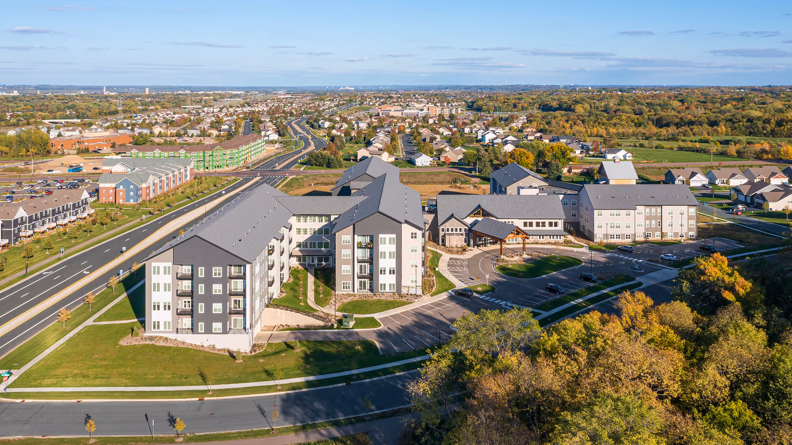 Aerial view of Benedictine Living Community- Shakopee Windermere Way, showing multiple large residential buildings with gray roofs and white and gray exteriors, surrounded by parking lots, roads, and green spaces. The surrounding area includes residential neighborhoods, trees, and a clear blue sky.