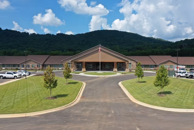 Front exterior view of Laurel Lodge Assisted Living & Memory Care building with a wide driveway, neatly trimmed grass, small trees, parked cars, and a mountainous backdrop under a partly cloudy sky.