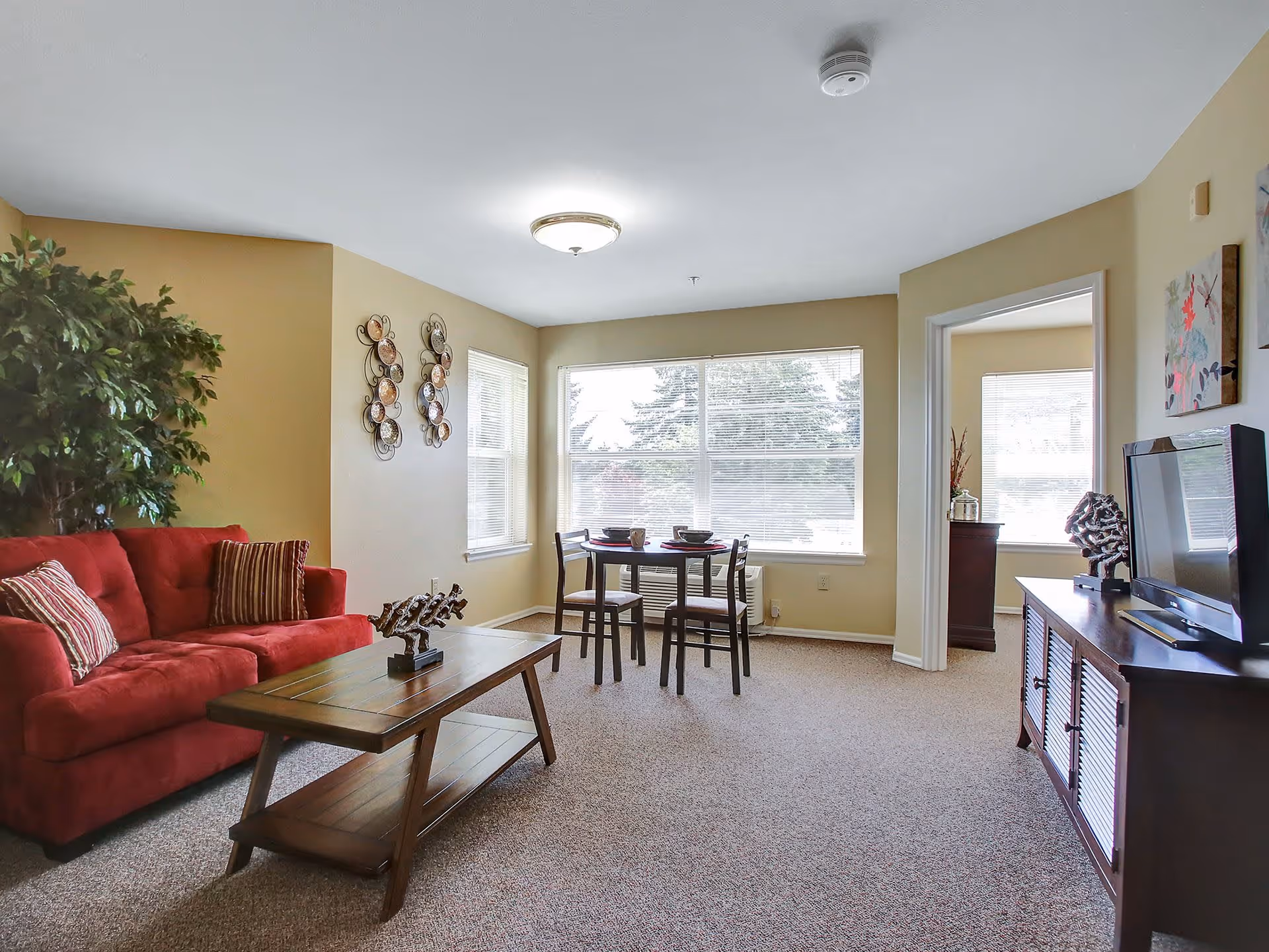 A bright living room with beige walls and carpeted floor featuring a red sofa with striped pillows, a wooden coffee table with a decorative sculpture, a small dining table with four chairs near large windows, and a wooden TV stand with a flat-screen television and decorative items.