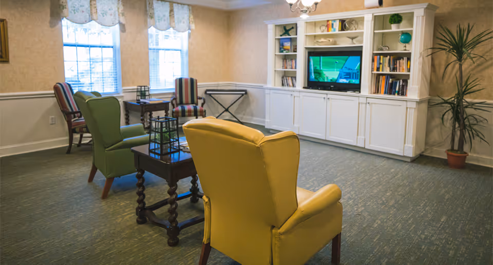 A cozy living room area with a yellow armchair and a green armchair facing a television set inside a white built-in bookshelf. There are two striped chairs near the windows with floral valances, a small wooden table with decorative lanterns, and a potted plant in the corner.