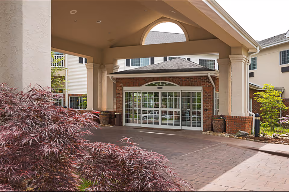 Entrance of Lake Pointe Landing Retirement Village showing a covered driveway with large pillars, a brick facade around automatic glass doors, and landscaped plants in the foreground.