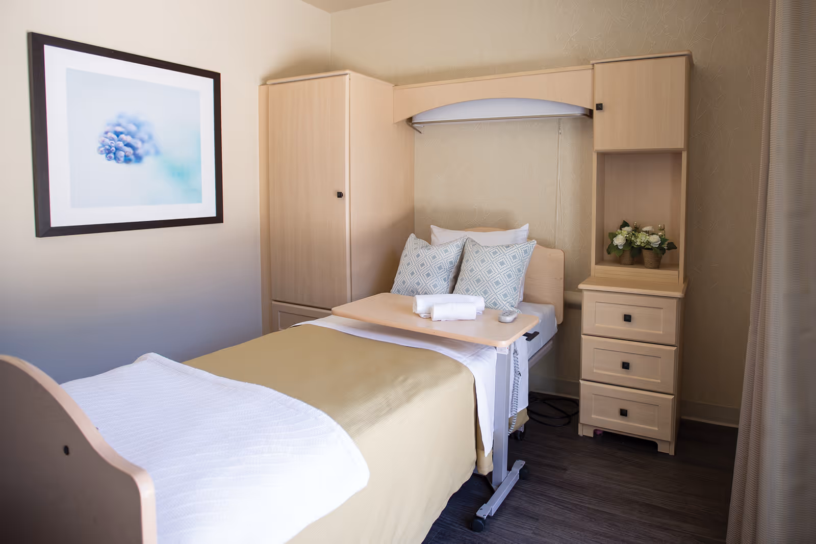 A neatly made single bed in a senior living facility room with beige bedding, two patterned pillows, and a white towel on a wooden overbed table. The room features light wood furniture including a wardrobe and a chest of drawers with a small plant on top. A framed picture of a close-up of a pine cone hangs on the wall.