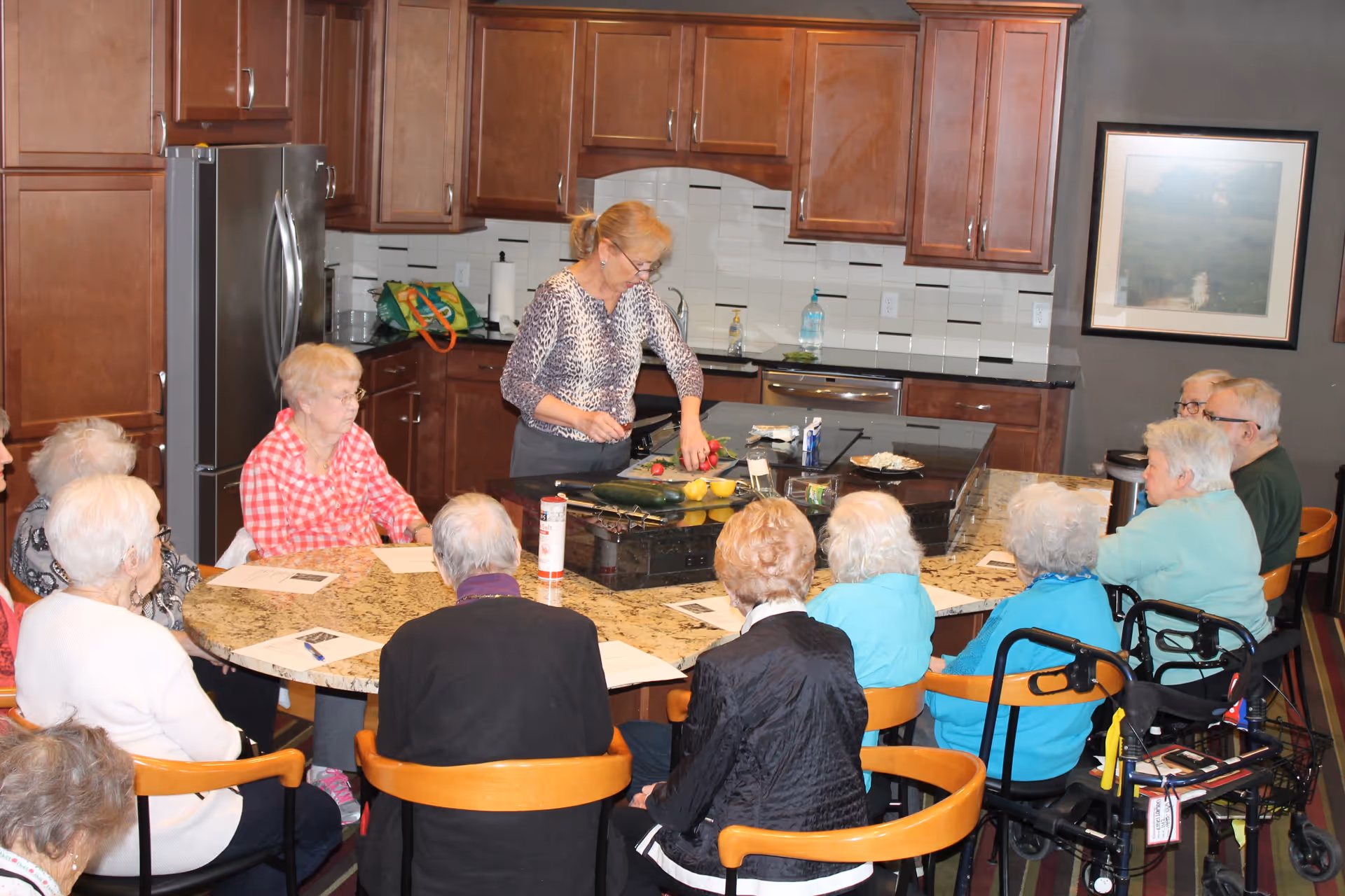 A group of elderly people seated around a large kitchen island in a senior living facility kitchen. A woman stands at the island demonstrating or preparing food while the seated individuals watch attentively. The kitchen features wooden cabinets, a stainless steel refrigerator, and a tiled backsplash.