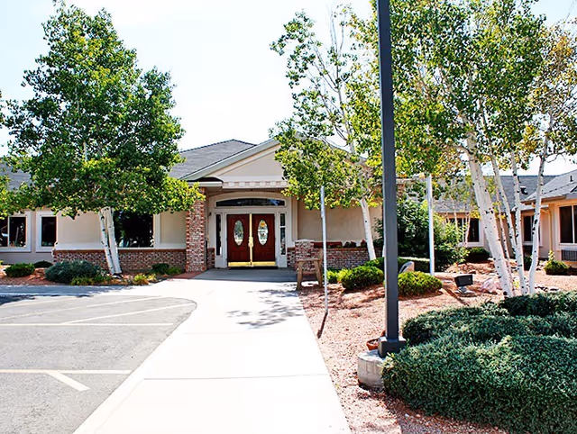 Sidewalk leading to the front entrance of an assisted living building with double doors, trees, and landscaped beds.