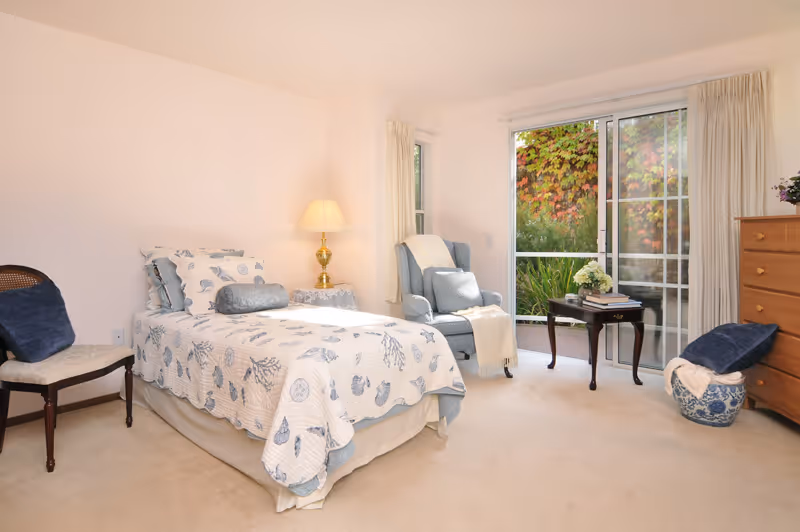 A bright bedroom with a single bed covered in a white and blue patterned quilt. Next to the bed is a small table with a lamp. There is a cushioned armchair with a throw blanket and pillow near a sliding glass door that opens to a garden with green and red foliage. A wooden dresser and a chair with a dark blue cushion are also in the room.