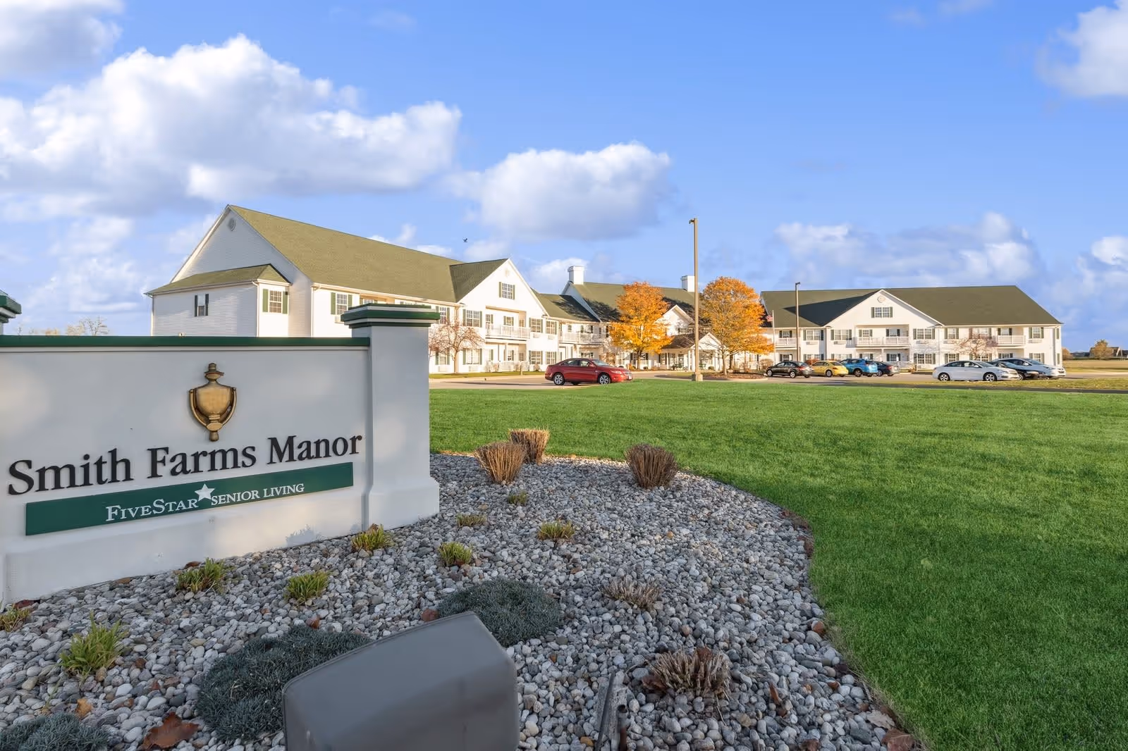 Exterior view of Smith Farms Manor senior living facility with a large green lawn, a stone-covered landscaped area in the foreground, and a white multi-story building with a green roof in the background under a partly cloudy sky.