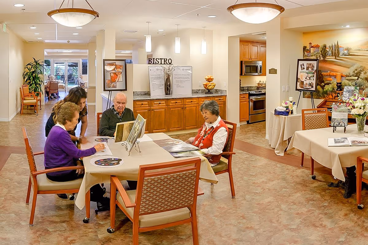 Three elderly individuals and a caregiver seated around a table engaged in an activity in a well-lit room with wooden cabinets, a kitchen area, and decorative artwork on the walls. The room has a warm, inviting atmosphere with tables covered in beige tablecloths and chairs with patterned upholstery.