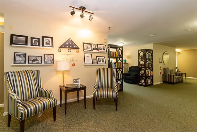 A cozy interior common area with two striped armchairs and a small wooden table with a lamp between them. The wall behind the chairs displays framed photographs and a folded American flag in a triangular case. In the background, there are bookshelves, a black recliner, and another seating area with a sofa and a round mirror on the wall. The space is warmly lit with ceiling lights and has carpeted flooring.