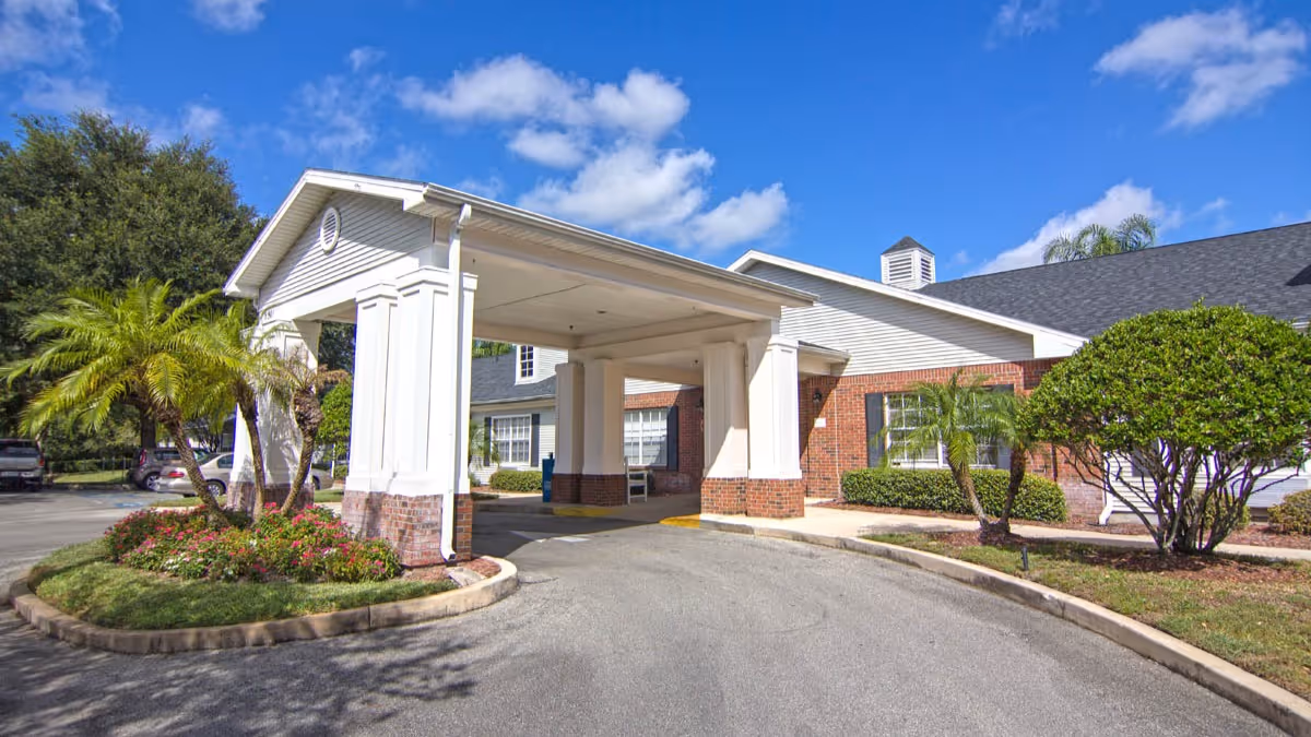 Exterior view of the entrance to a senior living facility with a covered drop-off area supported by white columns with brick bases. The building has white siding and brick accents, surrounded by landscaped greenery including palm trees and bushes under a blue sky with scattered clouds.