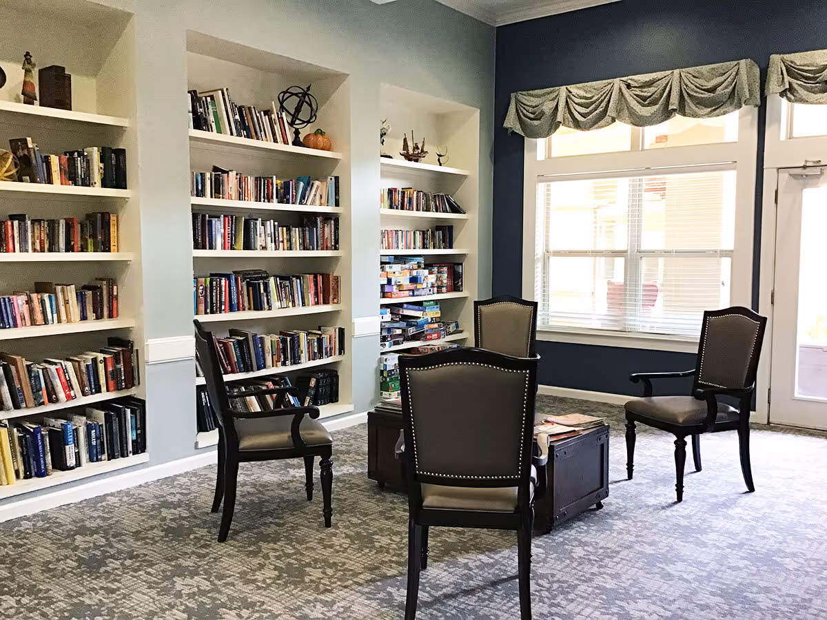 A cozy reading and game area in a senior living facility with built-in bookshelves filled with books and board games. Four cushioned chairs are arranged around a wooden coffee table on a patterned carpet. Large windows with valances allow natural light to brighten the room.