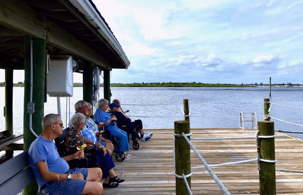 A group of elderly people sitting on benches under a covered wooden dock by the water, enjoying the view and holding drinks with lemon garnishes. The scene shows a calm body of water with a distant shoreline under a partly cloudy sky.