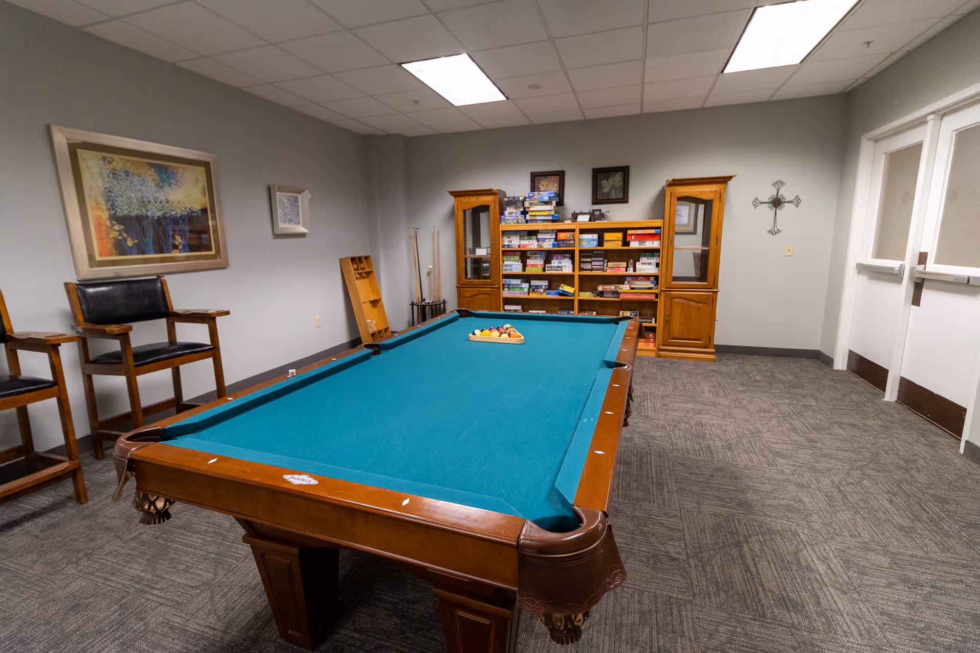 A game room with a pool table in the center, set up with billiard balls in a triangle rack. Behind the pool table, there is a wooden shelf filled with board games and two wooden cabinets on either side. Two tall chairs are positioned against the left wall, which is decorated with framed artwork. On the right wall, there is a decorative cross and double doors. The room has a carpeted floor and a drop ceiling with fluorescent lights.