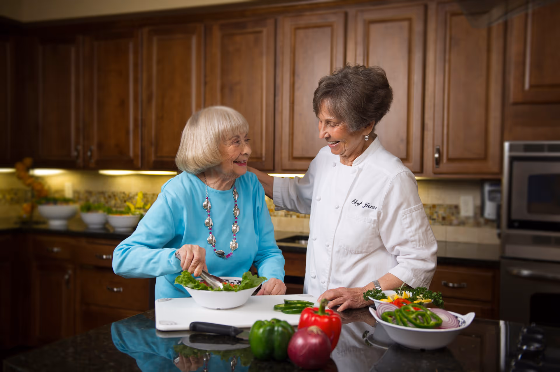 Two older women smiling and preparing a salad together at a kitchen island in a warm, wood-paneled kitchen.