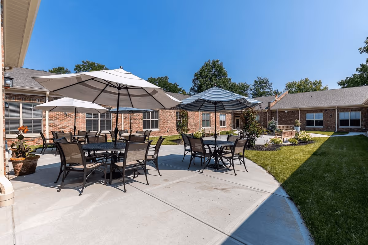 Outdoor patio area at The Victorian at Riverside featuring multiple round tables with chairs and large umbrellas providing shade. The patio is surrounded by a well-maintained lawn and brick buildings under a clear blue sky.