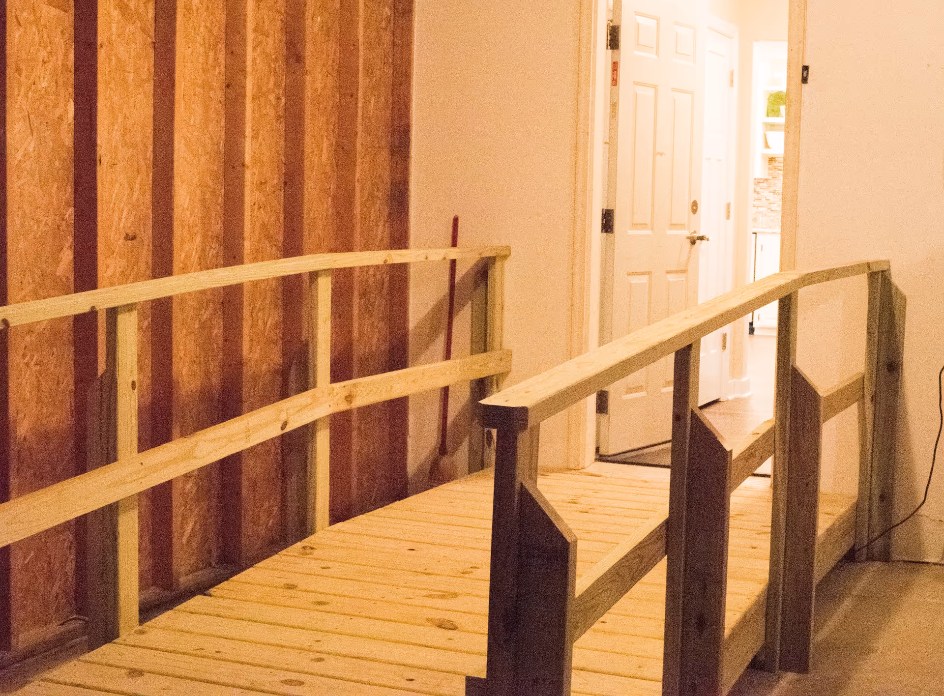 Interior view of a wooden ramp with handrails leading to a doorway inside a building. The walls are partially unfinished with exposed wooden studs on one side and painted drywall on the other. The doorway opens into a well-lit room with white doors and cabinetry.