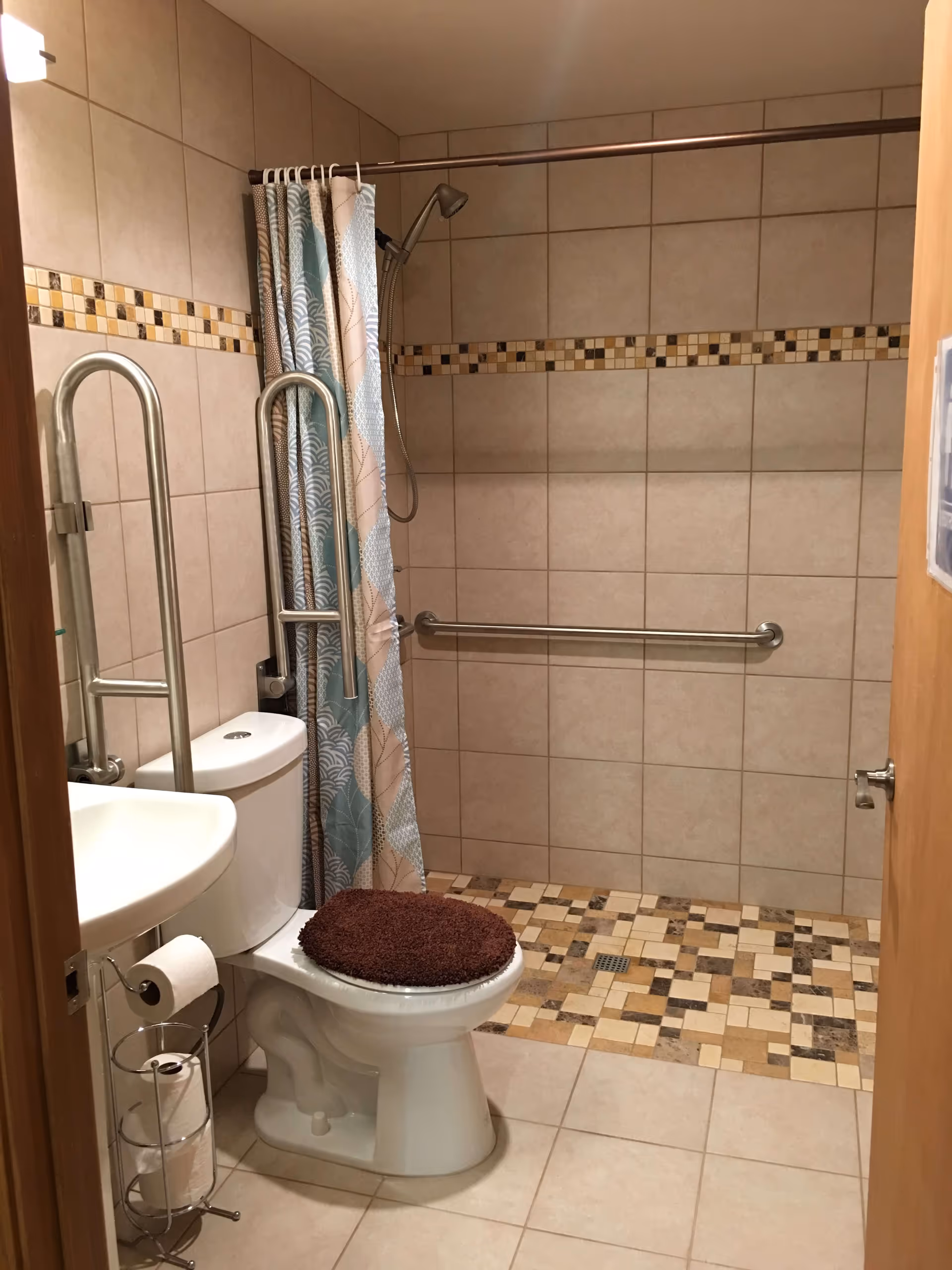 A bathroom with beige tiled walls and floor, featuring a toilet with a brown cushioned seat cover, a small white sink, a toilet paper holder with extra rolls, a shower area with a patterned shower curtain, and stainless steel grab bars for accessibility.