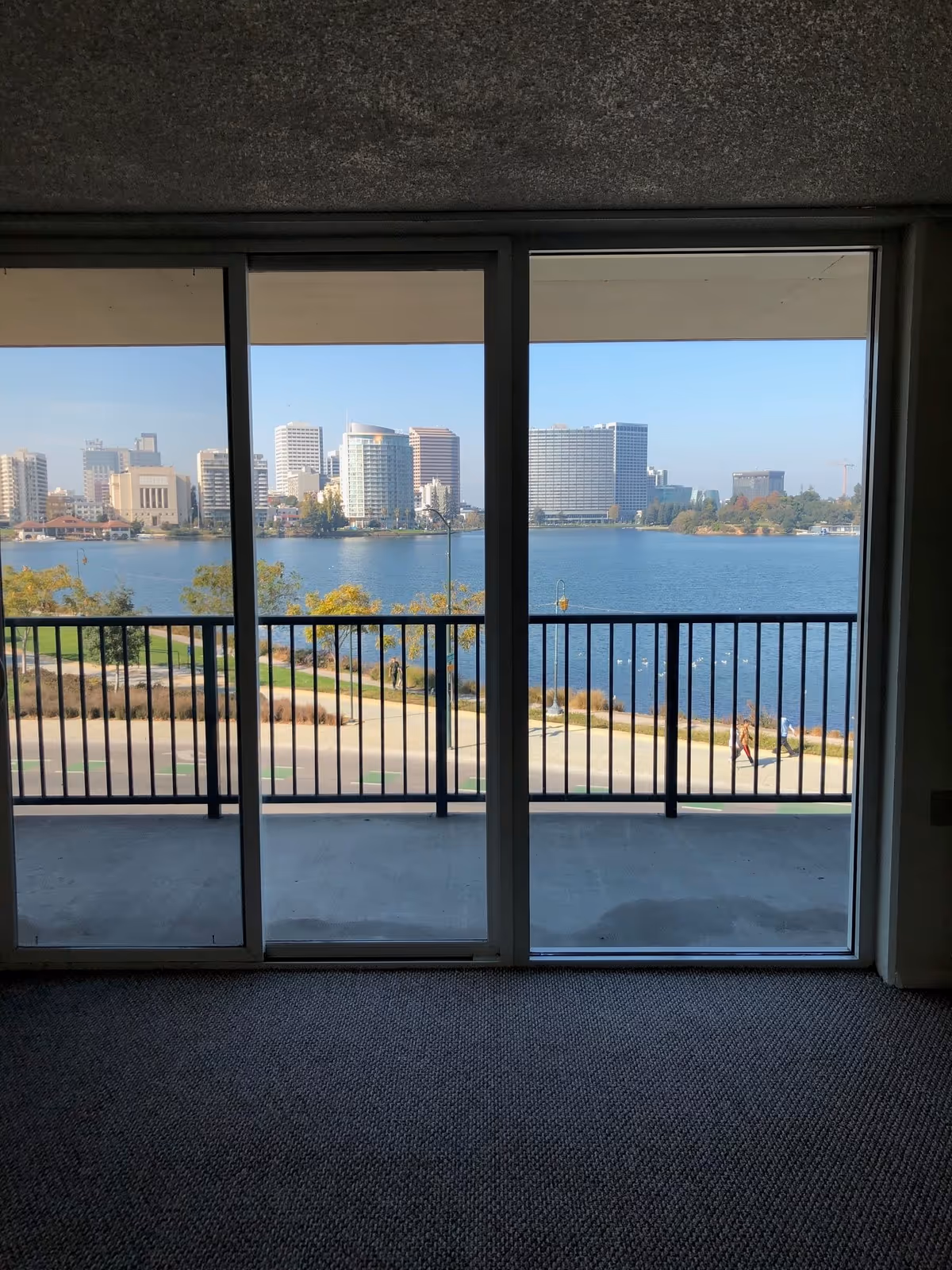 View through large sliding glass doors showing a balcony with black railing overlooking a body of water and a city skyline with multiple buildings under a clear blue sky.