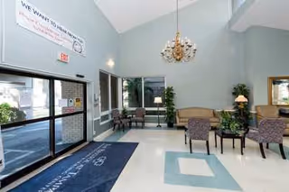 Lobby area of The Bristol at Tampa Rehabilitation & Nursing Center with seating arrangements including chairs and a sofa, a chandelier hanging from the ceiling, large windows, and glass entrance doors. There is a welcome mat on the floor and a banner on the wall above the entrance.