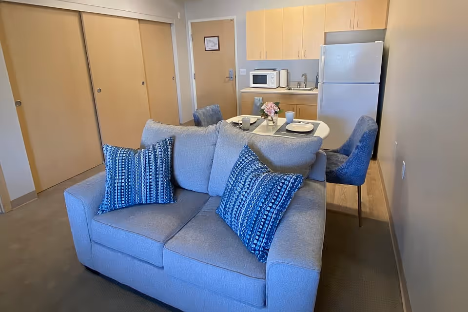 Small senior-living apartment interior with a gray loveseat and blue pillows facing a kitchenette and a small dining table with chairs.