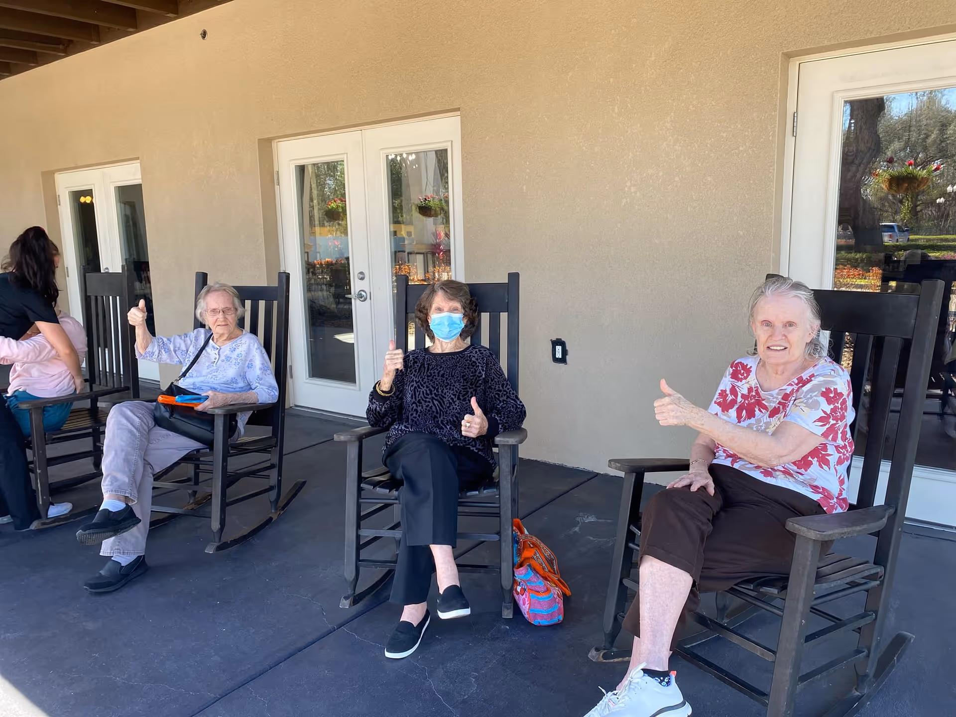 Three elderly women sitting on rocking chairs on a covered patio outside a building. Two women are giving thumbs up, and one woman is wearing a face mask. There is a woman in the background attending to a child. The patio has beige walls and glass doors.