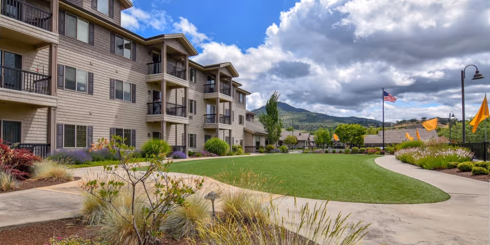Outdoor view of a senior living facility with a well-maintained lawn, landscaped garden beds, and a multi-story building with balconies. There are several orange flags and an American flag on flagpoles, with mountains and a partly cloudy sky in the background.