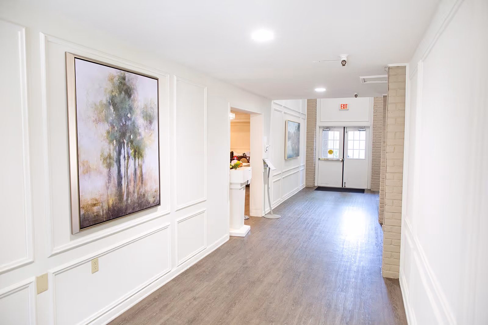 A bright hallway with white walls and wood flooring leading to a set of double doors with glass panels. The walls are decorated with framed paintings of trees, and there is a small stand with a sign near the entrance to another room on the left.