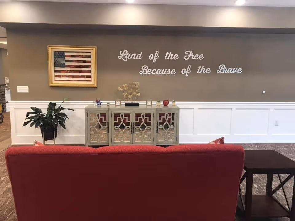 A communal seating area with a red couch facing a decorative console, an American flag painting, and wall lettering that reads 'Land of the Free Because of the Brave'.