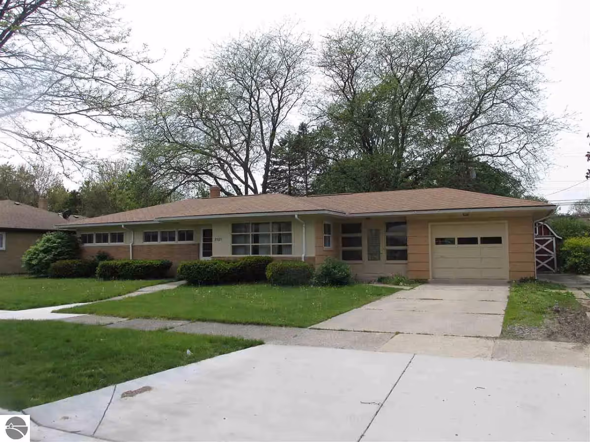 Single-story residential building with a front lawn, driveway, and attached garage. The house has a beige exterior with large windows and is surrounded by trees and shrubs.